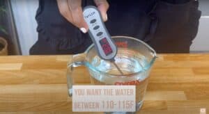 Digital thermometer measuring water temperature in a Pyrex cup, reads 110°F; ideal range is 110-115°F for recipes.